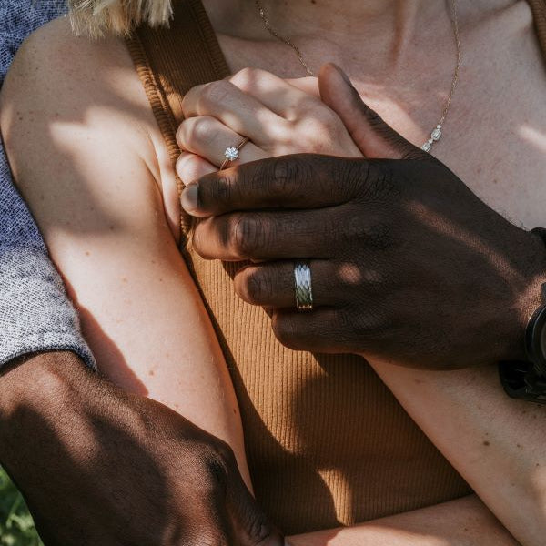 Close-up of two people holding hands with visible wedding rings, surrounded by greenery.